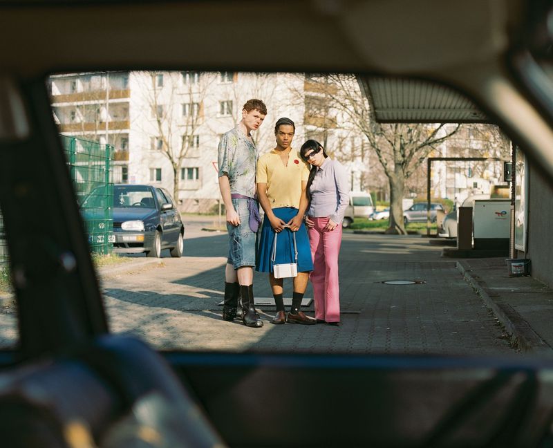 Three young adults pose outdoors near parked cars and buildings, viewed through a car window, showcasing casual street-style fashion on sunny day.