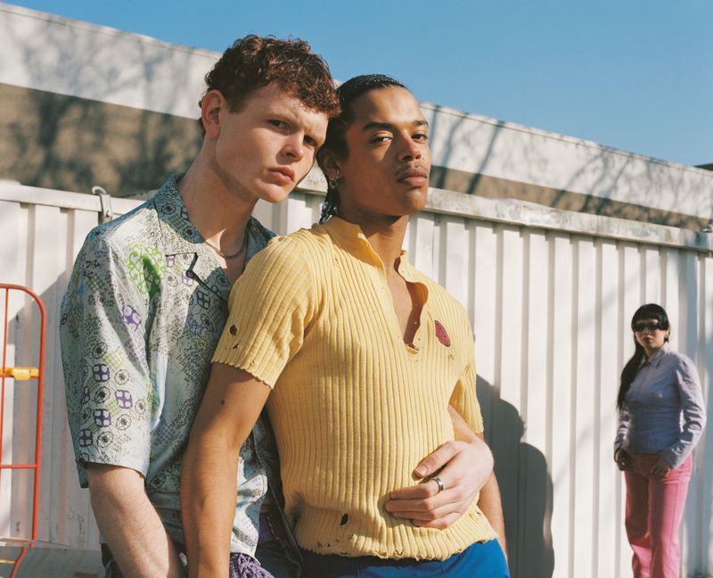 Two young men pose outdoors in casual shirts with a woman in the background, featuring urban fashion, bright daylight, and a container backdrop.