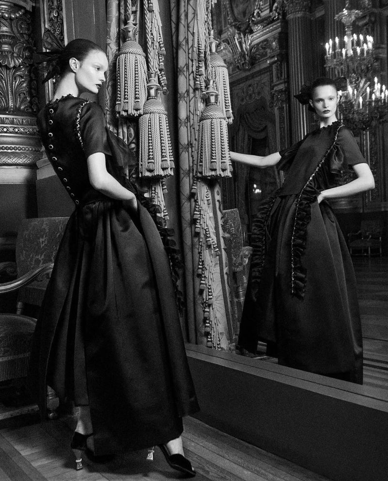 A woman in a black dress poses in front of a mirror inside an ornate, vintage room with decorative tassels, columns and chandelier.