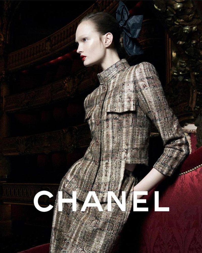 Fashion editorial shot of a model wearing a tweed Chanel skirt suit with a large bow in her hair, posing in front of a dark background.