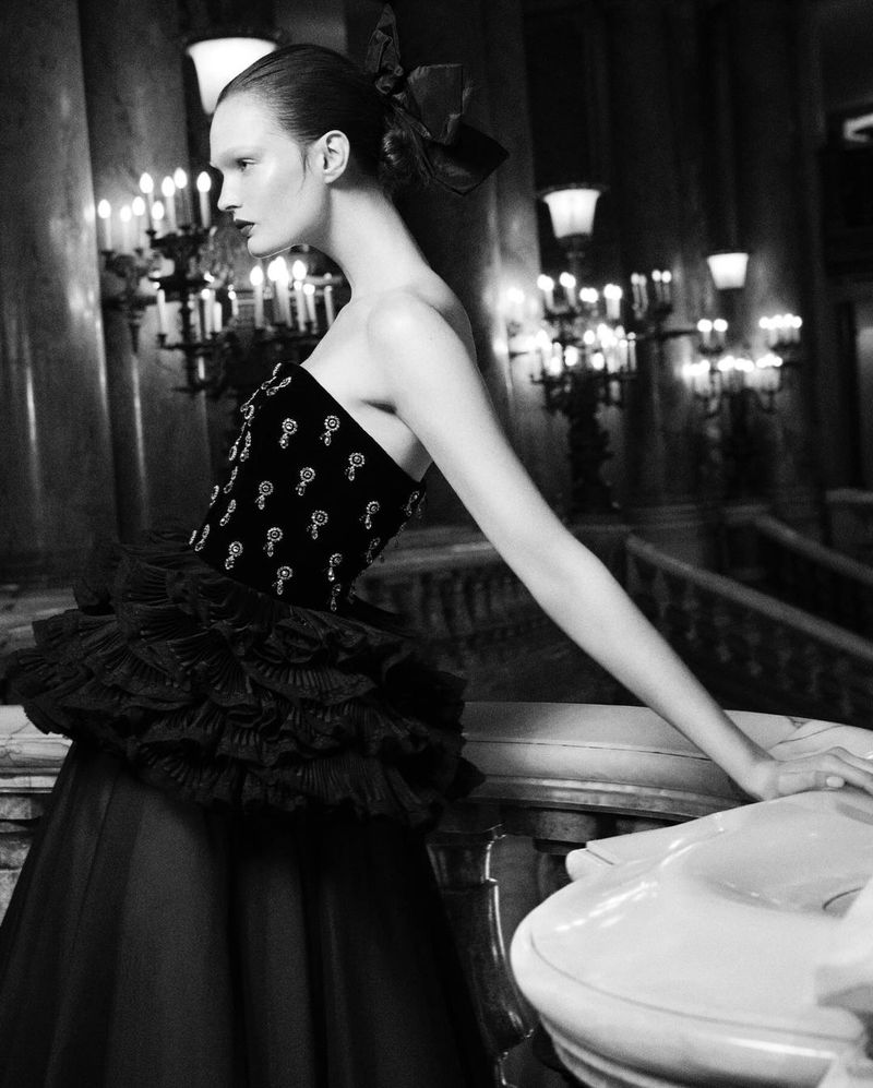 Black and white portrait of a fashion model in a dark dress with a bow in her hair standing in an ornate building with chandeliers.