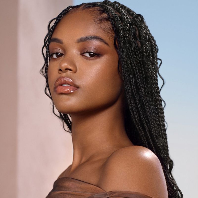 Portrait of a young black woman with braided hair, wearing a sheer brown top, posing against a light blue and beige background.