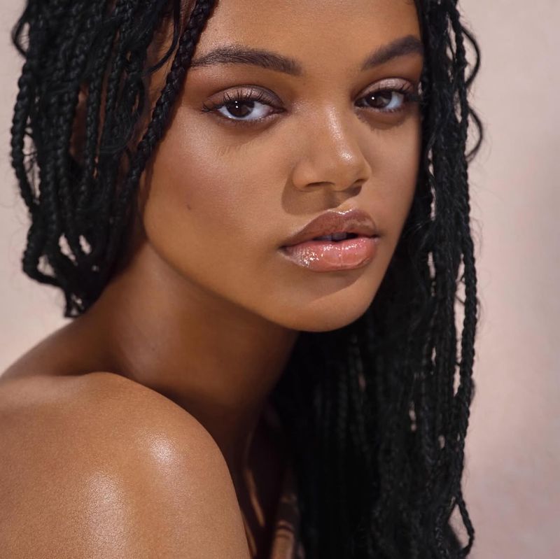 Close-up portrait of a young adult woman with braided hair, highlighted makeup, and a neutral expression against a light-pink background.