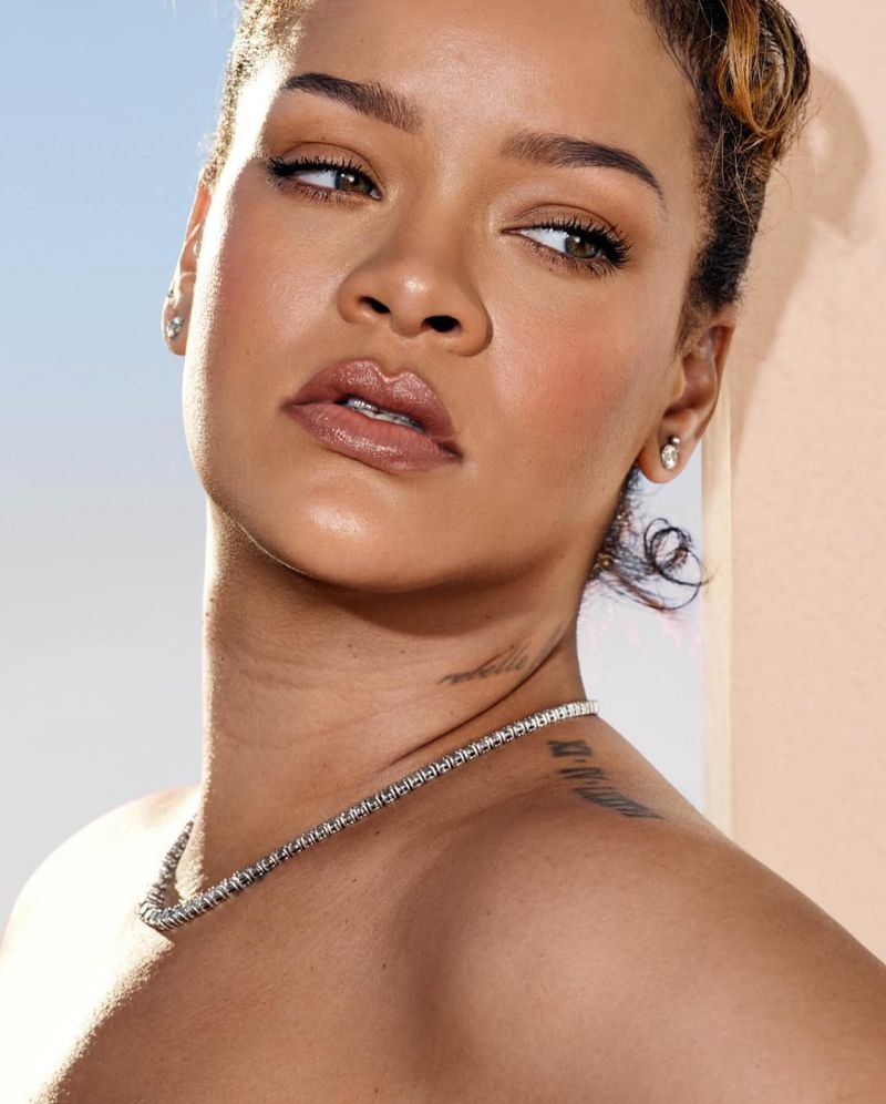 Close-up of Rihanna with tan skin wearing diamond necklace and stud earrings, set against light blue and peach background with soft lighting.
