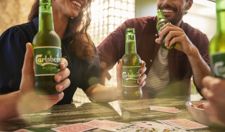Two adults sharing green-bottled Carlsberg pilsner beer, playing cards on a table. The pair are smiling, enjoying a friendly game indoors.