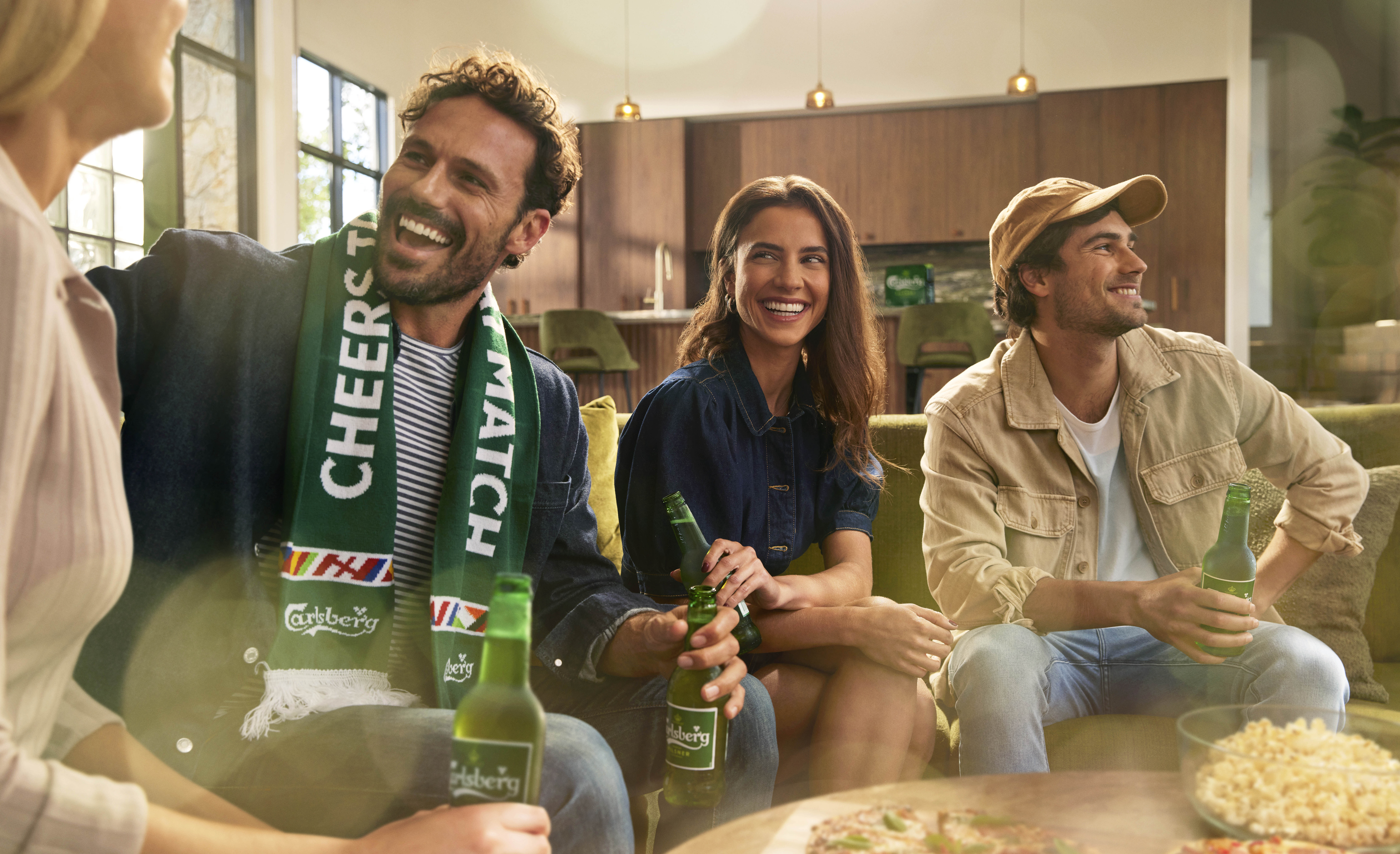 A group of four adults socializing indoors on a green sofa, holding beer bottles while watching a match and enjoying snacks.
