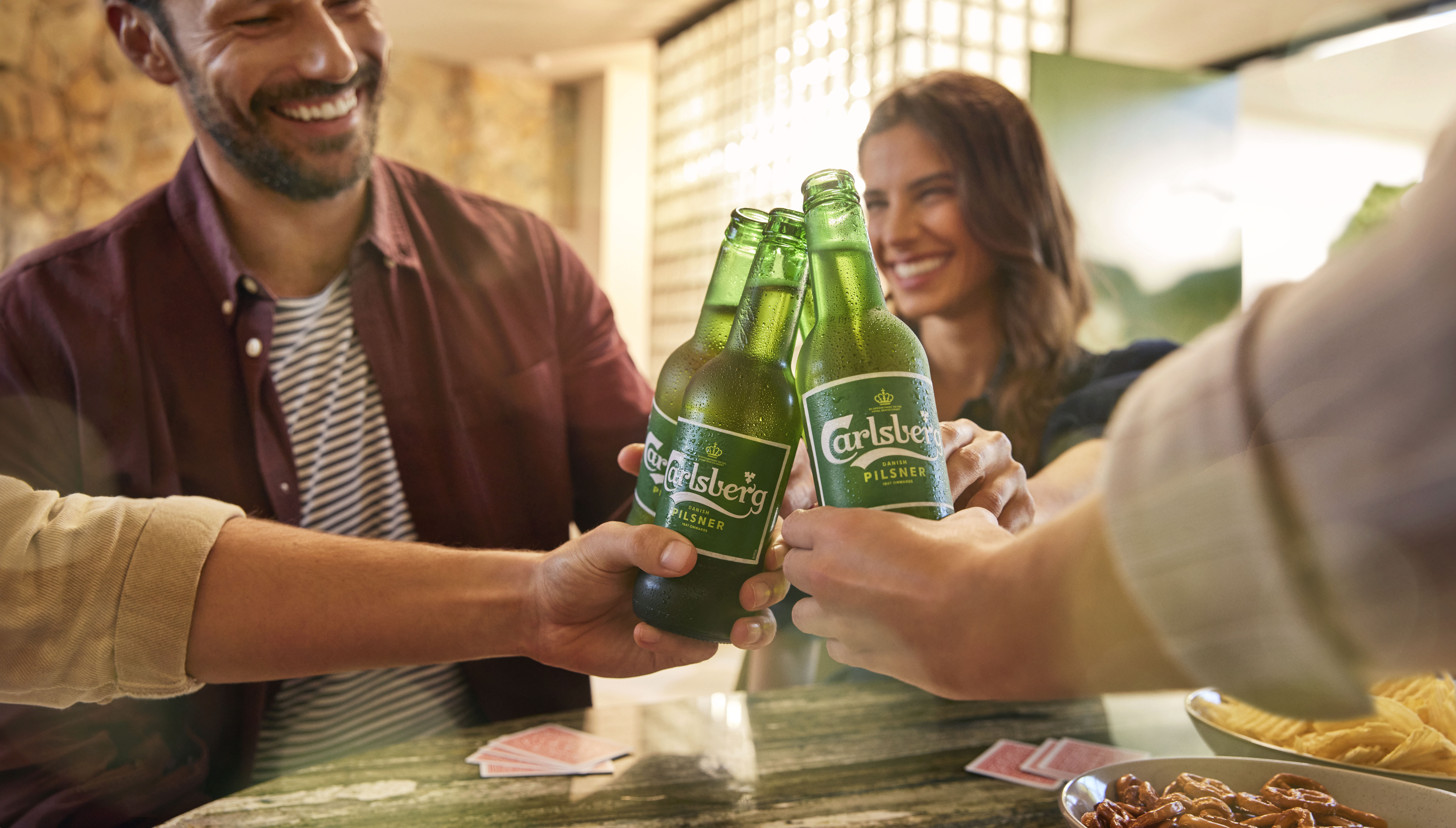 Four people cheers with green glass beer bottles, surrounded by snacks and playing cards on a table indoors, showcasing a casual gathering.