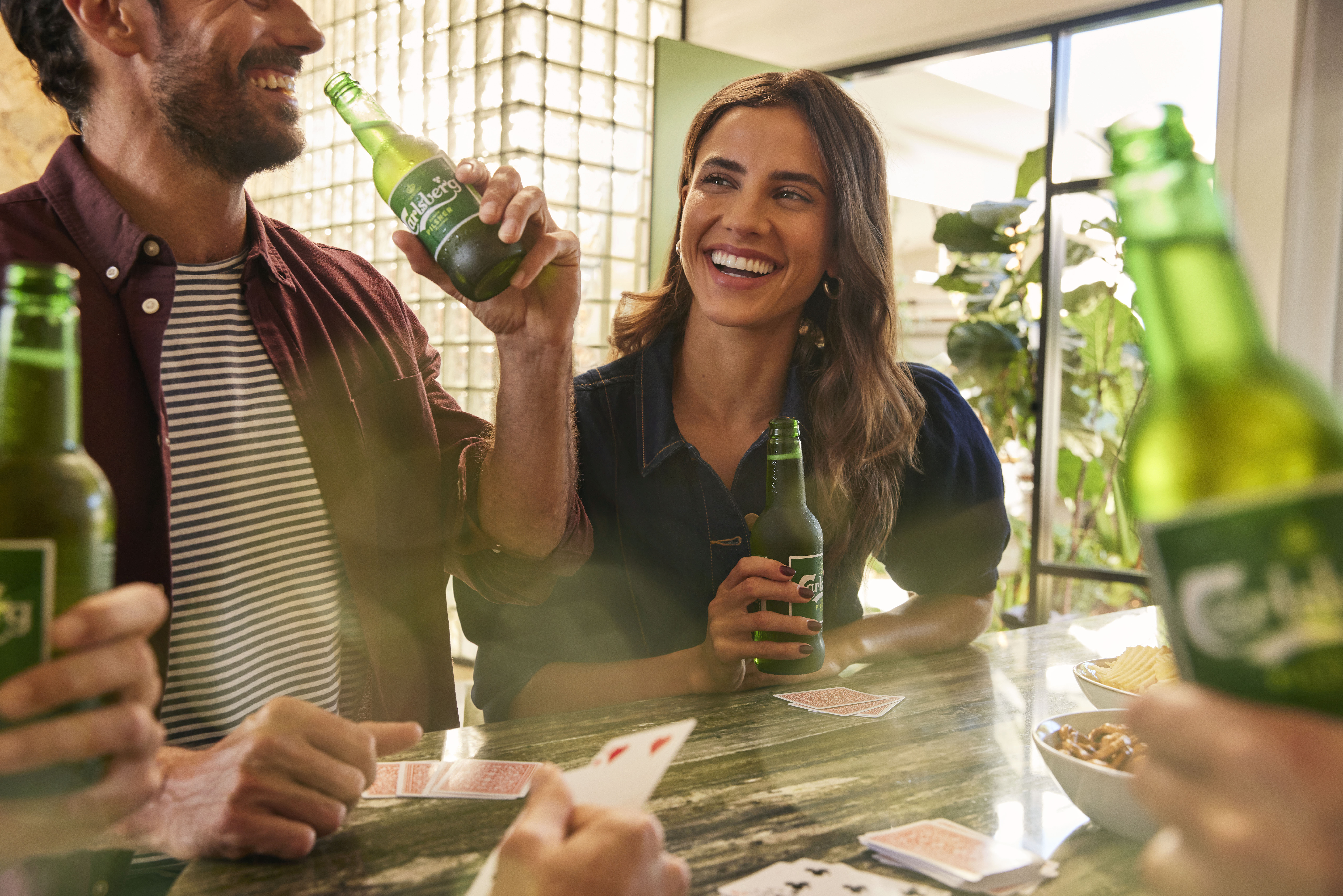 Group of friends laughing together while playing cards and drinking green bottled beer at a table indoors near a window.
