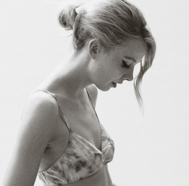 Monochrome portrait of a young woman with hair in a bun, gazing down with a tie-dye pattern bralette against a bright background.