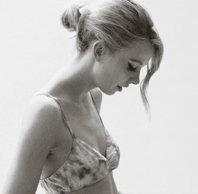 Monochrome portrait of a young woman with hair in a bun, gazing down with a tie-dye pattern bralette against a bright background.