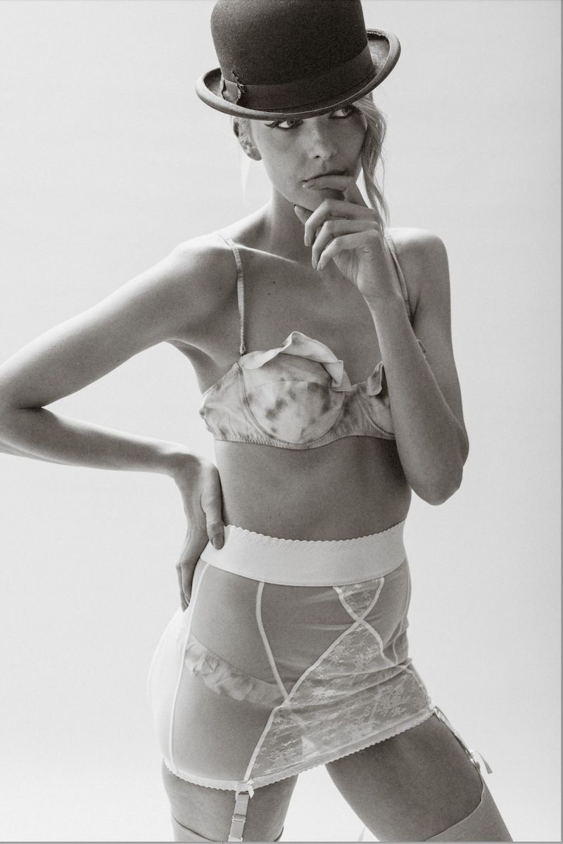 Studio shot of a woman wearing lingerie and a bowler hat. She is striking a pose with her hand on her chin in a bright environment.
