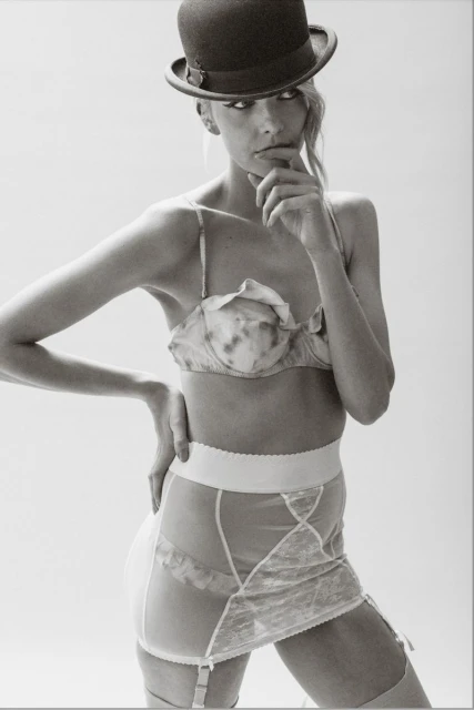 Studio shot of a woman wearing lingerie and a bowler hat. She is striking a pose with her hand on her chin in a bright environment.