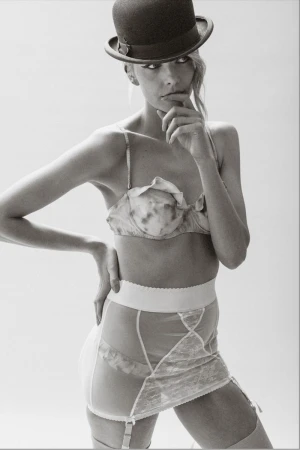 Studio shot of a woman wearing lingerie and a bowler hat. She is striking a pose with her hand on her chin in a bright environment.