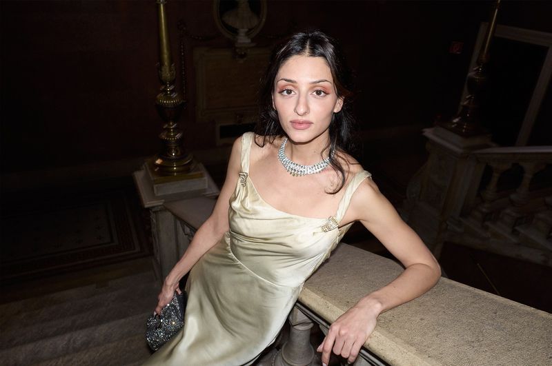 Elegant woman in satin dress and sparkling jewelry poses on marble stairs with ornate architectural details in the background.