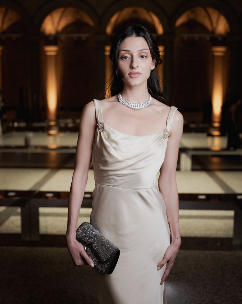 Elegant woman in gown and diamond necklace holds a sparkling clutch, posing in a stately hall with glowing sconces in the background.