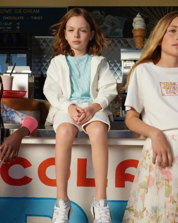 Three children sit on a white counter with vintage signage; two girls flank a central boy, all wearing casual summer clothing indoors.