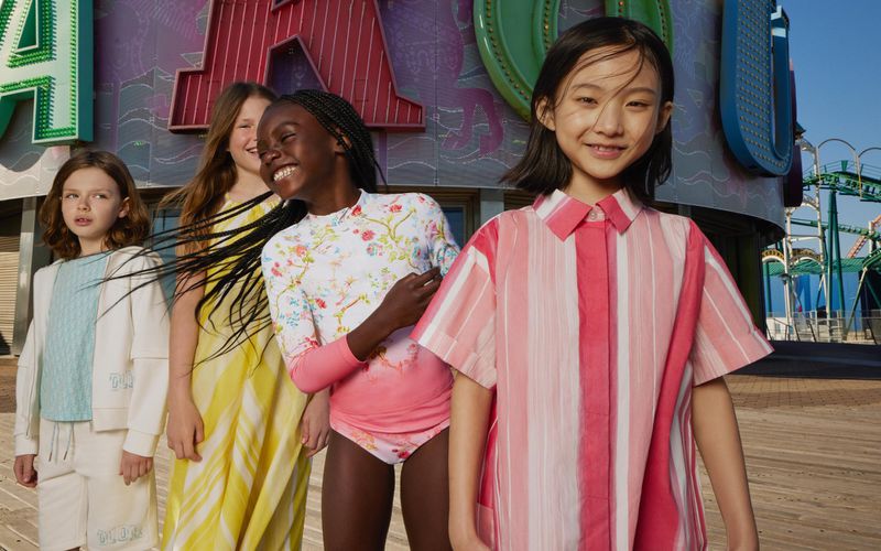 Four young girls stand smiling in front of an amusement park with a roller coaster. The girls are wearing fashionable clothing.