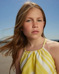 Close-up portrait of a child with long light-brown hair in a yellow-and-white striped halter dress against a clear blue sky.