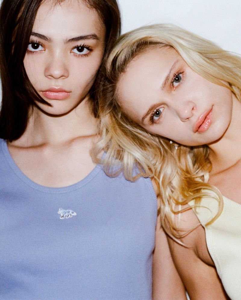 Two young women pose closely together in a studio shot, one with brunette hair in a blue top, the other blonde in yellow.