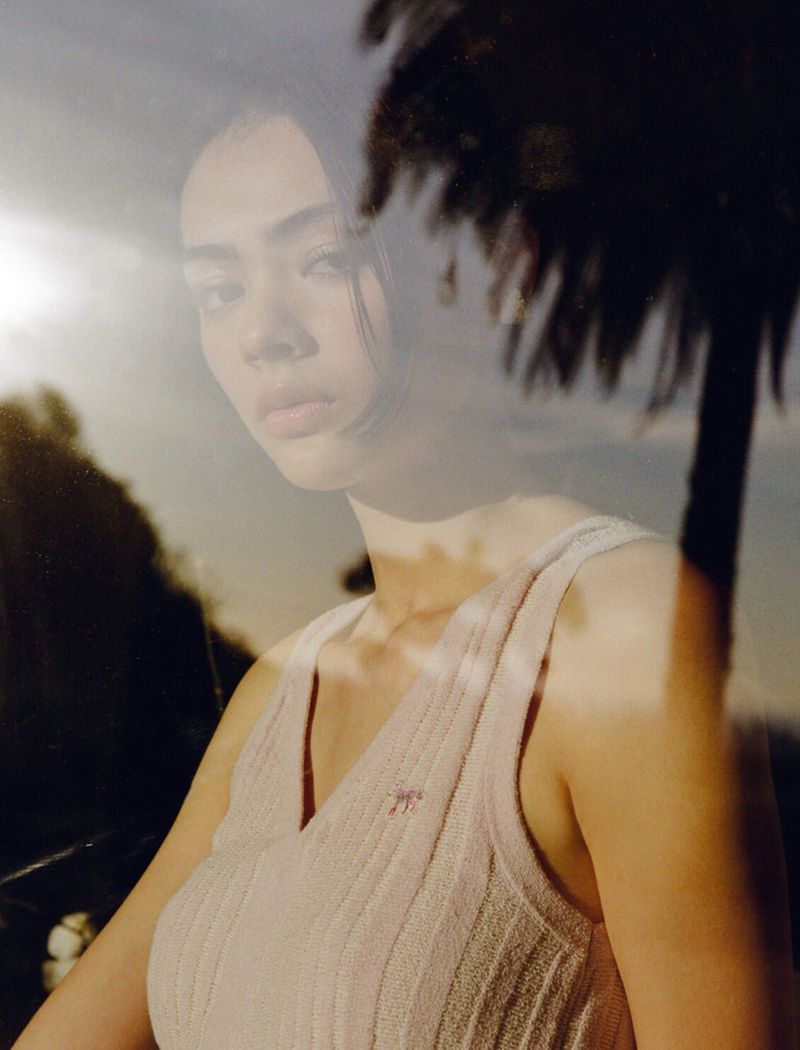 A young woman with dark hair poses outdoors, gazing slightly left, wearing a light-pink ribbed tank top with a blurred palm-tree backdrop.