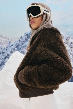 Portrait of an adult female wearing goggles and a brown sherpa jacket, posing in a snowy mountain landscape on an overcast day.