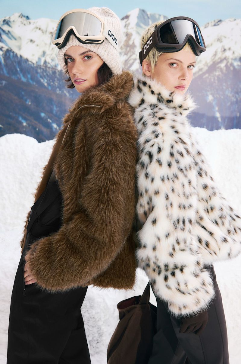 Two women wearing ski goggles and fur coats pose in a winter scene with snow and mountains, showcasing fashion and winter sports apparel.