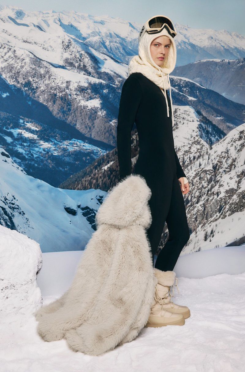 A woman in a black catsuit, ski goggles, and fur stole poses against a backdrop of snow-capped mountains in a full shot.
