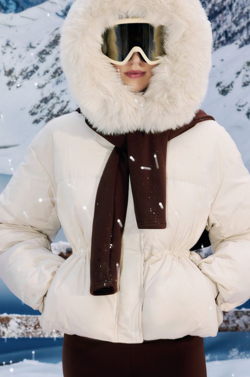 Woman in a white hooded puffer jacket, ski goggles, and brown scarf against a backdrop of snow-covered mountains and snowy conditions.