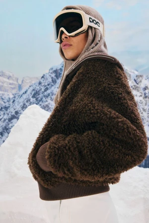Woman wearing a brown textured fleece jacket, knit balaclava hood, and white ski goggles against snowy mountain peaks.