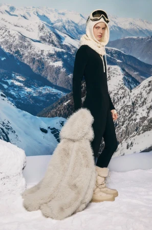 Model wearing black ski suit, cream balaclava and snow boots holding furry coat against mountain snowfield backdrop for Oysho