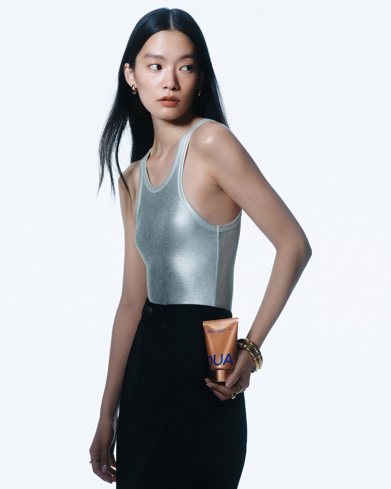 Attractive Asian woman posing in shiny silver tank top, black skirt, and gold jewelry, holding a tan bottle on white seamless backdrop.