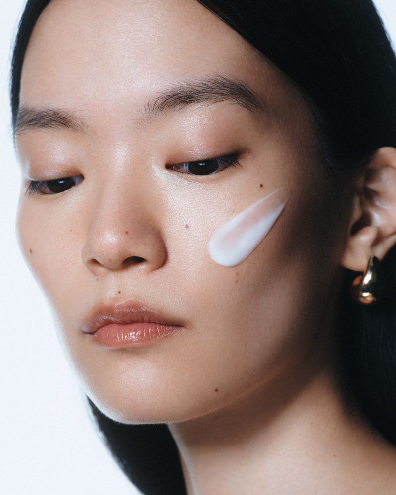 Close-up beauty portrait of an East Asian woman with a white skincare cream swatch on her cheek and a small gold earring.