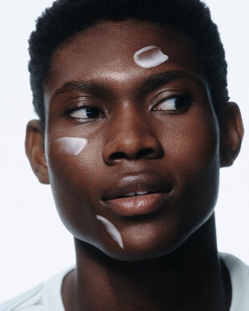 Close-up studio shot of an adult male model with dark skin and facial moisturizer on a white background for a beauty advertisement.