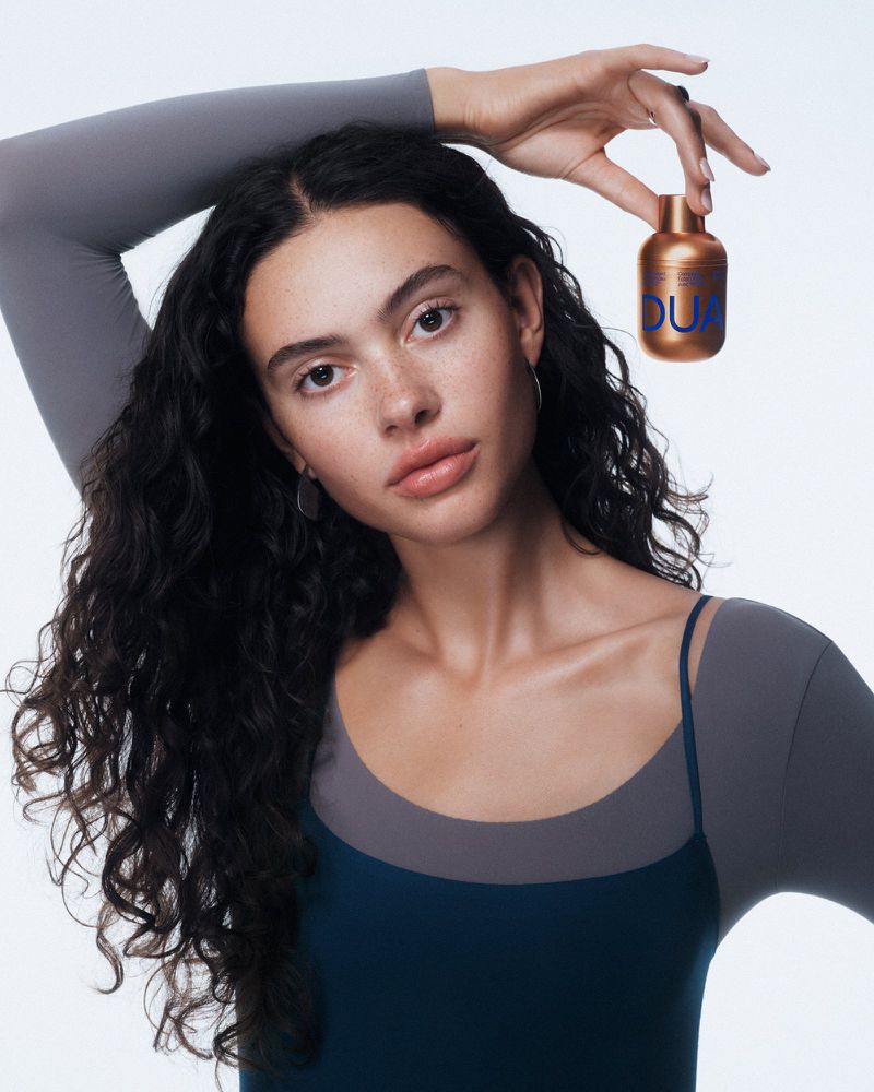 A young woman with long curly dark hair holds a gold bottle of Dua perfume, posing in front of a white backdrop. Beauty shot for advertising.