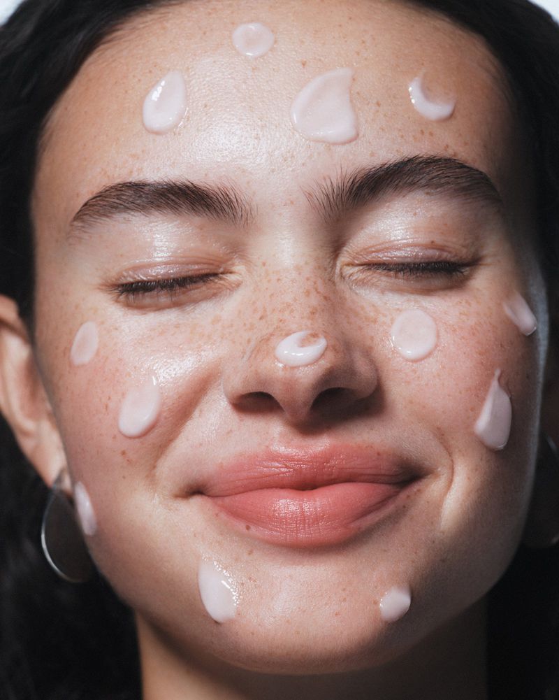 Close-up of a smiling fair-skinned woman with closed eyes and freckles. Light pink cream dots her face in a beauty skincare treatment.