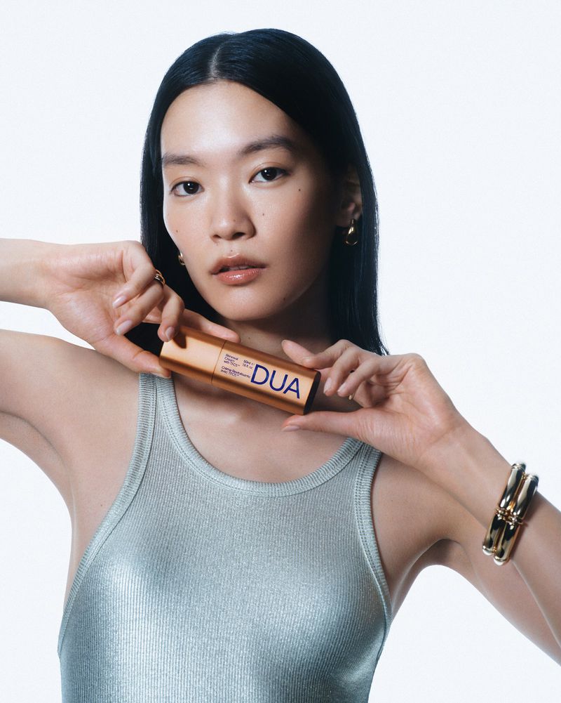 Close-up of an Asian woman in a tank top holding a DUA beauty product. Shot in a bright photo studio with a minimalist aesthetic.