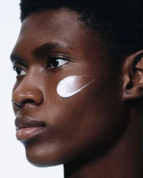 Close-up profile of a Black man with a white skincare cream swatch across his cheek against a plain white background.
