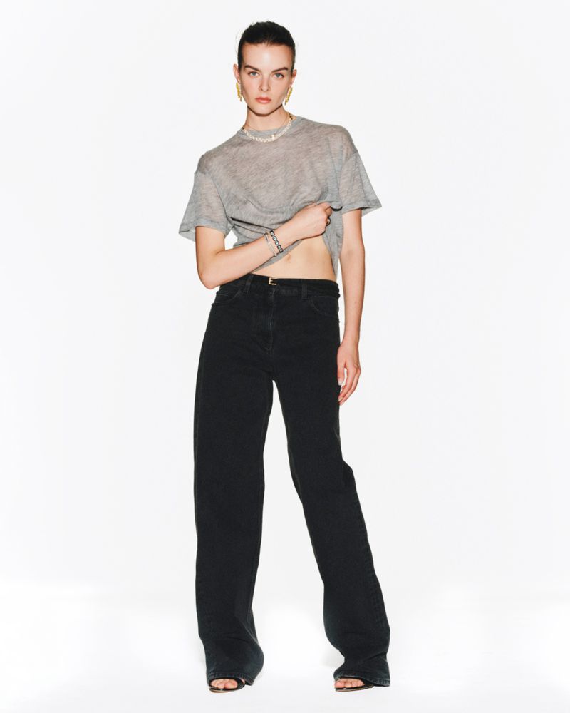 Fashion shot of a woman in a gray shirt and black wide-leg jeans standing in a brightly lit studio, looking at the camera.