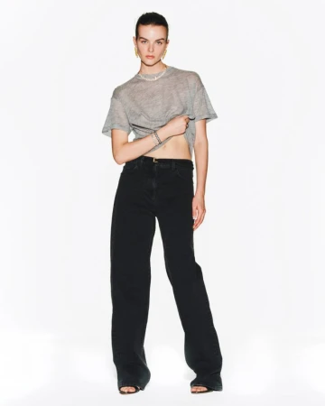 Fashion shot of a woman in a gray shirt and black wide-leg jeans standing in a brightly lit studio, looking at the camera.
