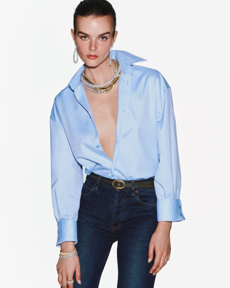 A woman poses in a light blue button-down shirt with dark denim jeans, accessorized with gold jewelry and a belt, against a white backdrop.