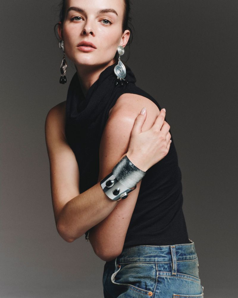 Portrait of an adult woman wearing a black turtleneck, metal cuff, and earrings against a neutral background in a fashion-editorial style.