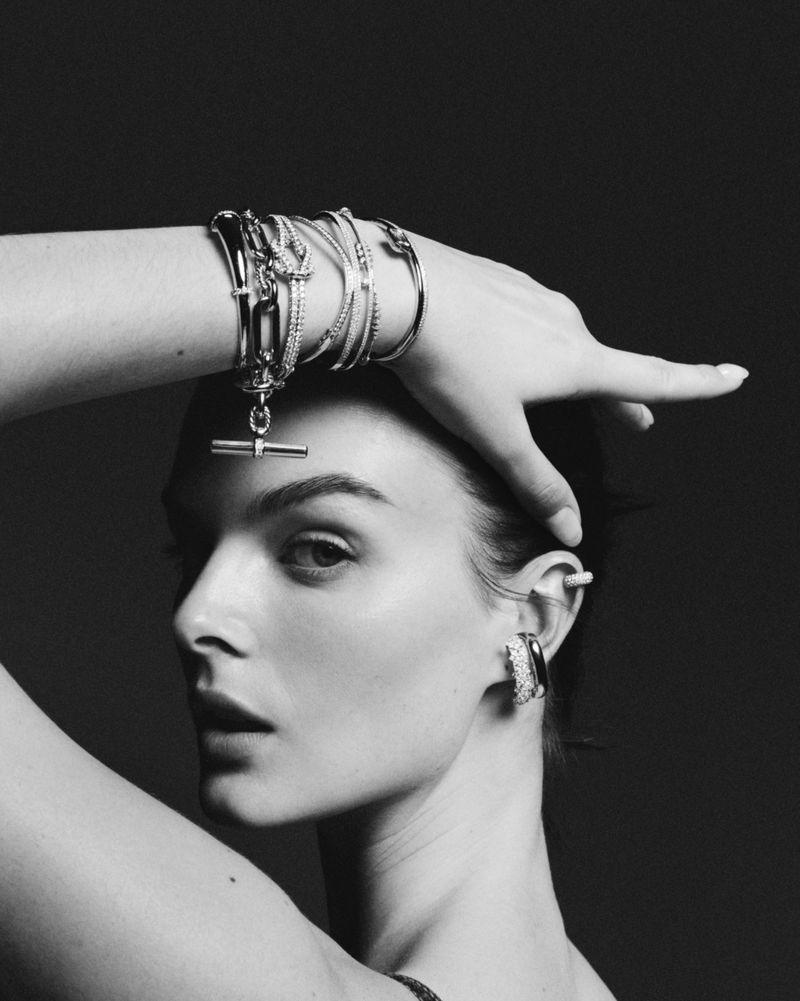 Black and white portrait of model Aylah Peterson posing with stacked metallic bracelets and ear jewelry against a dark background.