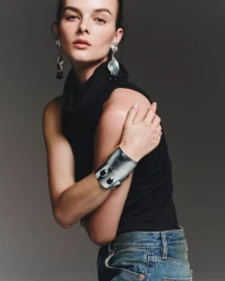 Aylah Peterson in a black sleeveless top and blue denim jeans wearing large silver earrings and a cuff bracelet against a grey background.