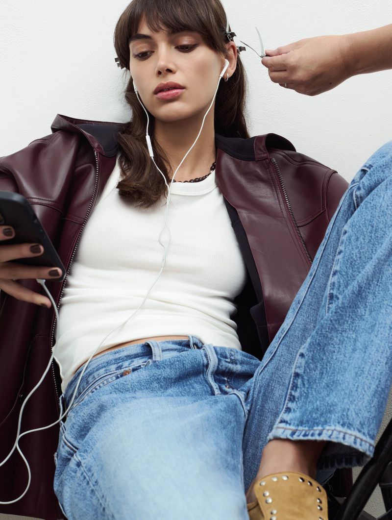 Young woman in a jacket sits wearing earphones and holding a smartphone while another hand adjusts her hair. 