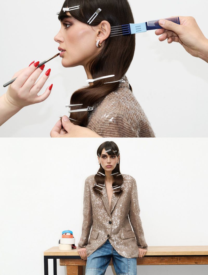 Brunette woman in a shimmering beige blazer and jeans, posing for a fashion shoot while a makeup artist applies lipstick.