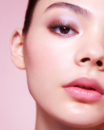 Close-up of a woman's face with fair skin, pink glossy lips, and mauve eye makeup. The background is light pink and soft lighting.