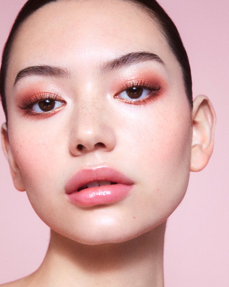 Close-up of a woman's face with pink eyeshadow and glossy lips against a plain pink backdrop in a beauty and makeup shot.