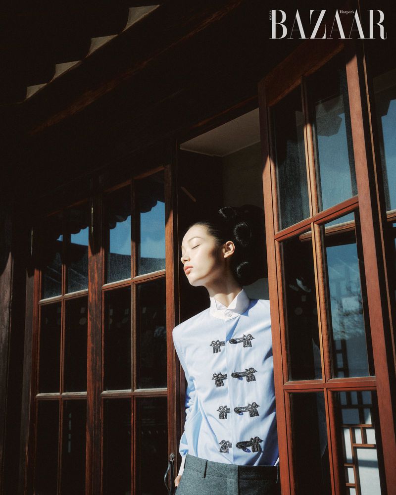 An Asian woman stands in an open doorway with a blue button-down shirt, illuminated by natural light. Editorial fashion shot for Bazaar magazine.
