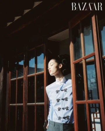 An Asian woman stands in an open doorway with a blue button-down shirt, illuminated by natural light. Editorial fashion shot for Bazaar magazine.
