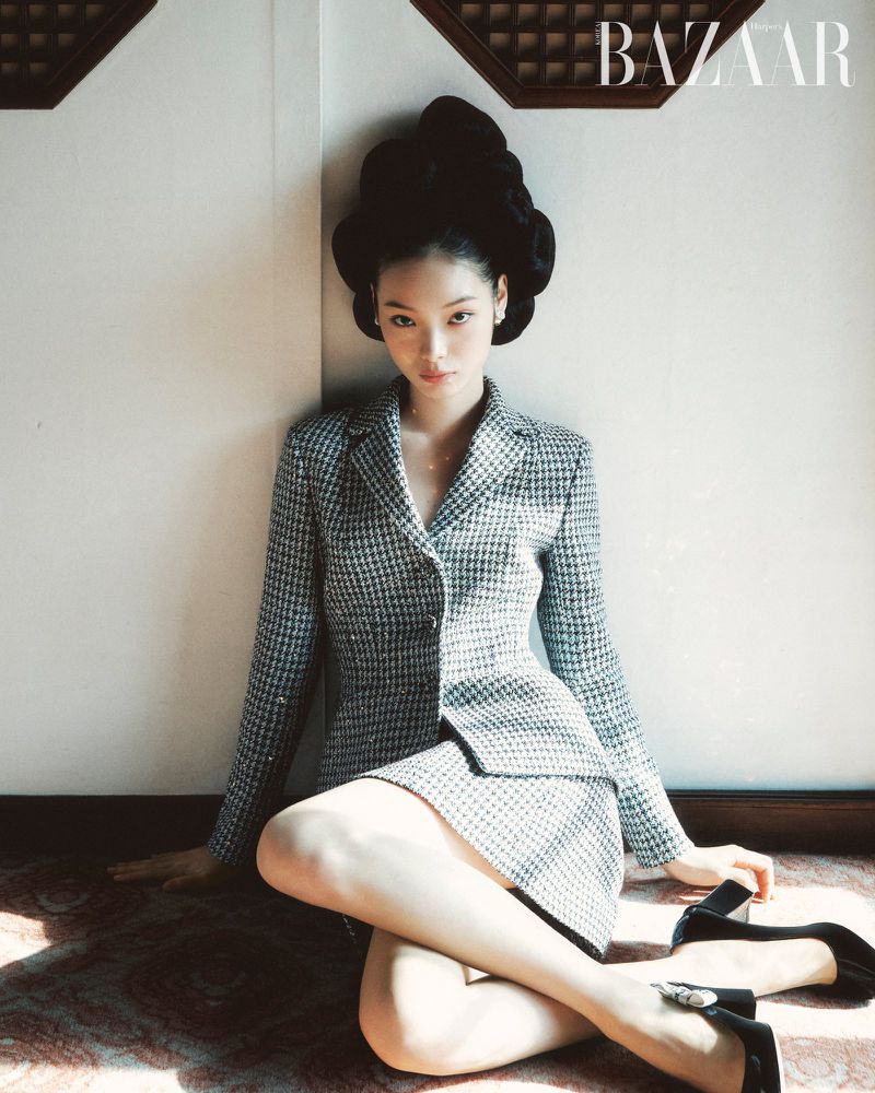 Elegant woman in a houndstooth blazer and skirt poses indoors with sculptural hair, accessorized with black pumps in a fashion editorial.